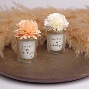 May include: Two small glass candles with floral accents. The candles have the text "I CAN'T SAY 'I DO' without you" printed on them. One candle has a peach-colored flower, and the other has a white flower. They sit on a brown plate.