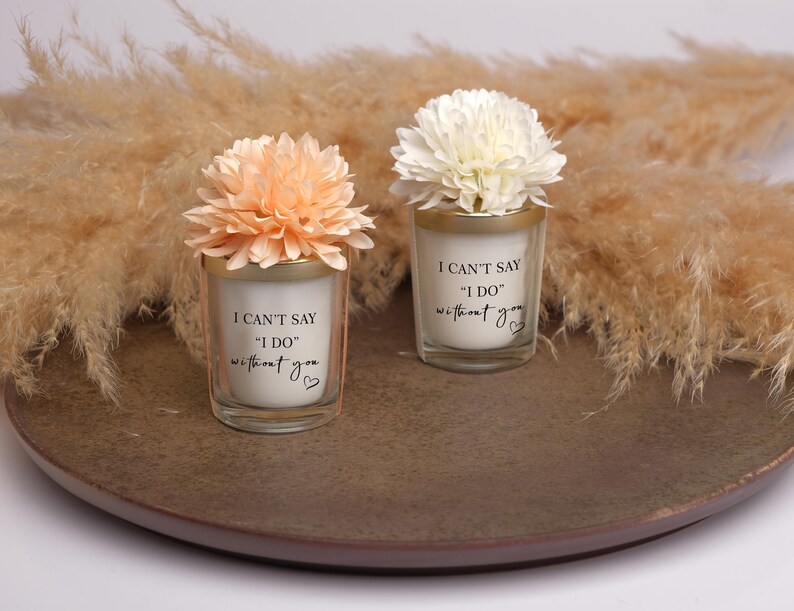 May include: Two small glass candles with floral accents. The candles have the text "I CAN'T SAY 'I DO' without you" printed on them. One candle has a peach-colored flower, and the other has a white flower. They sit on a brown plate.