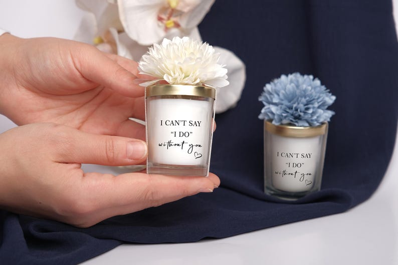 May include: Two small, clear glass candles with gold rims and floral accents. The candles have the text "I CAN'T SAY 'I DO' without you" printed on them. One candle has a white flower, the other a blue flower.