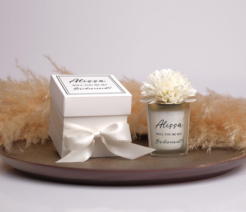May include: A white gift box tied with a satin bow, and a candle in a glass holder, both with the text "Alissa, Will You Be My Bridesmaid?" A white flower sits atop the candle. The items are on a brown tray.
