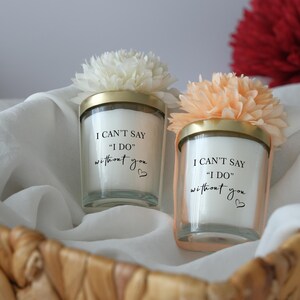May include: Two glass candles with gold lids and floral accents, inscribed with "I CAN'T SAY 'I DO' without you." The candles sit in a woven basket with white fabric and red flowers in the background.