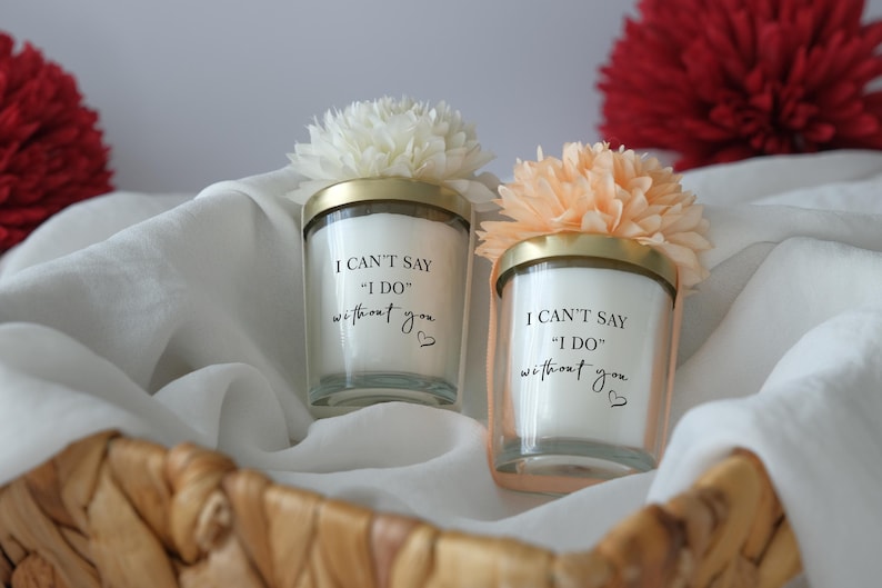 May include: Two glass candles with gold lids and floral accents, inscribed with "I CAN'T SAY 'I DO' without you." The candles sit in a woven basket with white fabric and red flowers in the background.