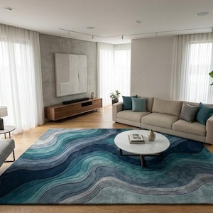 May include: A modern living room with a large blue and teal wave-patterned rug. The room features a beige sofa with blue pillows, a round marble coffee table, and a gray armchair. Large windows with white curtains provide natural light.