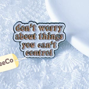 May include: A metal enamel pin featuring a white background with a black border and the brown text "don't worry about things you can't control" in a rounded font. A small cream-colored tag with the text "CraftsBeeCo" is attached to the pin.