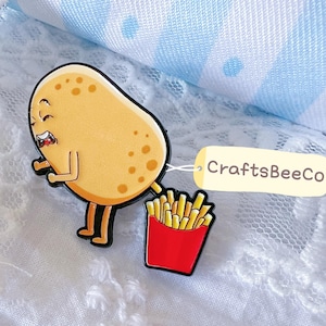 May include: A tan potato character enamel pin with a cartoon face and brown spots, depicted in a bending position. A red container filled with yellow french fries is attached to the potato. A small cream-coloured tag reads CraftsBeeCo.
