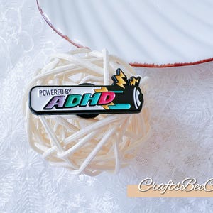 Op de afbeelding: Een zwart-witte pin met de tekst "POWERED BY ADHD" in kleurrijke letters. De pin heeft een bliksemschicht en een batterijontwerp. De pin is geplaatst op een bol wit touw.