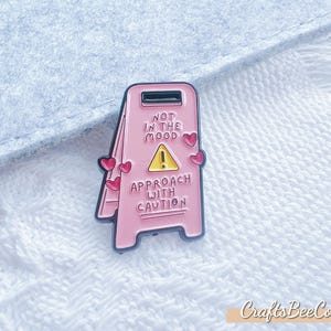 Könnte beinhalten: Ein rosa Emaille-Pin in Form eines Warnschildes mit dem Text „NOT IN THE MOOD“ und „APPROACH WITH CAUTION“. Das Schild hat ein gelbes Dreieck und rote Herzakzente. Der Pin befindet sich auf einer weißen, strukturierten Oberfläche.