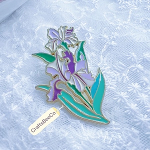 Könnte beinhalten: Ein goldfarbener Metall-Irisblumen-Anstecknadel mit lila, weißen und grünen Emaille-Details. Die Anstecknadel hat ein detailliertes Design der Blütenblätter und Blätter der Blume. Das Etikett an der Anstecknadel lautet CraftsBeeCo.