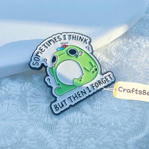 Op de afbeelding: Een groene kikker-vormige pin met de tekst "Sometimes I Think But Then I Forget." De pin heeft een witte buik, een kleine hoed en vraagtekens. Een klein label met de tekst "CraftsBeeCo" is bevestigd.