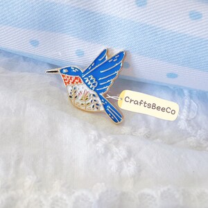 May include: A hummingbird enamel pin featuring a vibrant blue body, a bright orange throat patch, and intricate white floral patterns on the chest. The pin is attached to a small, light-coloured tag with the text CraftsBeeCo printed in black.