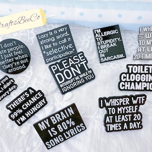 May include: A collection of ten black enamel pins with white text featuring humorous phrases such as I don't hate people, Lazy is a very strong word, I'm allergic to stupidity, Please don't interrupt me, Toilet clogging champion, and My brain is 80% song lyrics.