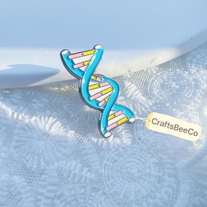 May include: A blue, pink, and yellow DNA double helix pin with a silver outline. The pin has a tag that reads "CraftsBeeCo". The pin is on a white surface.