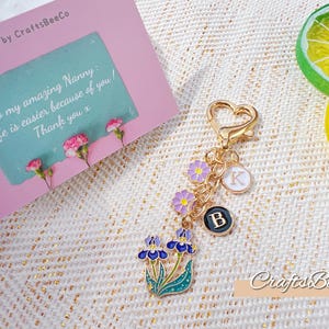 Puede incluir: Un llavero dorado con cierre en forma de corazón, adornado con dijes florales y colgantes con iniciales. El llavero presenta un dije de flor de iris azul y morado, junto con una tarjeta que dice "To my amazing Nanny: Life is easier because of you! Thank you x."