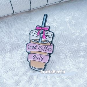 May include: Enamel pin depicting an iced coffee cup, adorned with a pink bow and straw. A pink banner on the pin reads "Iced Coffee Girly". The pin is set against a white lace background.