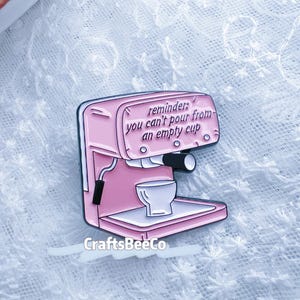 Könnte beinhalten: Ein rosa Emaille-Pin in Form einer Espressomaschine. Der Pin hat den Text "reminder: you can't pour from an empty cup". Er hat eine weiße Tasse und einen schwarzen Griff. Der Markenname "CraftsBeeCo" steht unten.