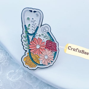 Könnte beinhalten: Ein floraler Stethoskop-Anstecker mit türkisfarbenem Umriss. Der Anstecker zeigt ein Stethoskop-Design, das mit rosa, roten und gelben Blumen und grünen Blättern gefüllt ist. Ein Etikett mit dem Text "CraftsBeeCo" ist angebracht.