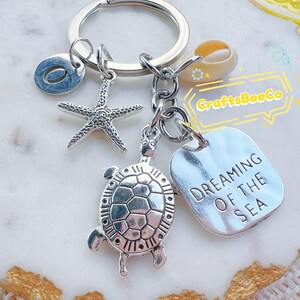 Op de afbeelding: Zilverkleurige sleutelhanger met zee-thema bedels. Inclusief een zeester, schildpad, schelp en een tag met de tekst "DREAMING OF THE SEA". De sleutelhanger heeft ook een letter "O" bedel.