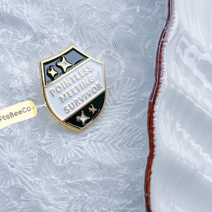 May include: A shield-shaped enamel pin with the text "Pointless Meeting Survivor." The pin has a gold border, black and white sections, and star accents. A small tag with "CraftsBeeCo" is attached.