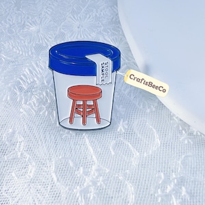 May include: A novelty pin featuring a clear plastic container with a blue lid, a red stool graphic, and a label that reads "STOOL SAMPLE." A yellow tag with "CraftsBeeCo" is attached.