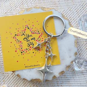 May include: A silver star keychain with a ring, hanging from a yellow card with the text "You are my star". The card has a star design and the text "CraftsBeeCo". The keychain is on a decorative surface.