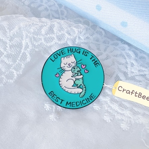 Könnte beinhalten: Ein runder, blaugrüner Emaille-Pin mit einer weißen Katze, die eine grüne Eidechse mit rosa Herzen umarmt. Der Text lautet LOVE HUG IS THE BEST MEDICINE. Ein kleines cremefarbenes Etikett am Pin zeigt den Text CraftBeeCo in schwarzer Schrift.