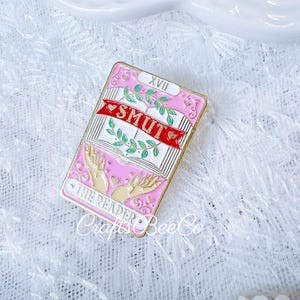 May include: Pink and gold enamel pin with a tarot card design. The pin features the word "SMUT" on a banner, with "THE READER" at the bottom. The pin has a pink background and a gold border.