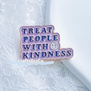 Könnte beinhalten: Eine Metall-Emaille-Anstecknadel mit hellviolettem Hintergrund und dunkelblauer Prägeschrift, die TREAT PEOPLE WITH KINDNESS lautet. Die Anstecknadel enthält ein kleines weißes, kreisförmiges Smiley-Symbol, das neben dem Wort WITH integriert ist.