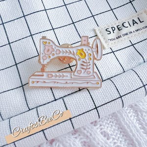 May include: A white and pink enamel sewing machine pin with floral details and gold accents. The pin is on a white fabric with a black grid pattern. A small tag reads "SPECIAL" and "YOU ARE ONE IN A MILLION".