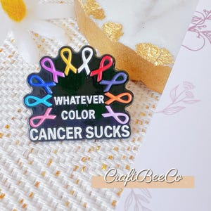 Peut inclure: Épinglette en émail noir avec des rubans de sensibilisation colorés et le texte "WHATEVER COLOR CANCER SUCKS". L'épinglette présente des rubans bleus, roses, violets, jaunes, rouges et oranges. L'épinglette est sur une surface texturée.