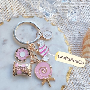 Könnte beinhalten: Ein goldfarbener Schlüsselanhänger mit mehreren Anhängern. Zu den Anhängern gehören ein rosa Donut, ein rosa-weißes Bonbon, ein rosa Schokoriegel mit dem Wort "sweet", ein rosa Lutscher und ein Anhänger mit dem Buchstaben "F". Der Schlüsselanhänger hat ein Etikett mit der Aufschrift "CraftsBeeCo".
