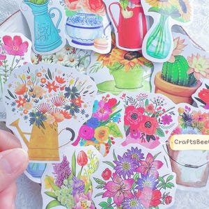 Puede incluir: Una colección de pegatinas florales coloridas con varios arreglos florales en jarrones, regaderas y macetas. Las pegatinas muestran una variedad de flores en colores vibrantes, incluyendo rosa, rojo, amarillo y morado. Las pegatinas están sobre un plato blanco.