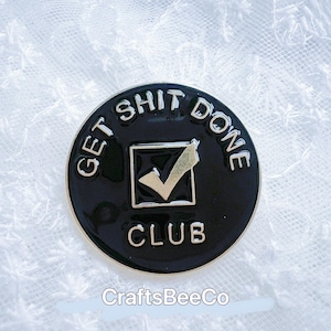 Op de afbeelding: Een zwarte en gouden ronde pin met de tekst "GET SHIT DONE CLUB". De pin heeft een gouden vinkje in een vierkant. De pin ligt op een wit, getextureerd oppervlak.