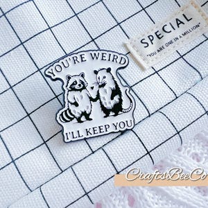 May include: A black and white enamel pin depicting a raccoon and an opossum holding hands. The pin reads "YOU'RE WEIRD I'LL KEEP YOU". A small tag with the word "SPECIAL" and the text "YOU ARE ONE IN A MILLION" is also visible.