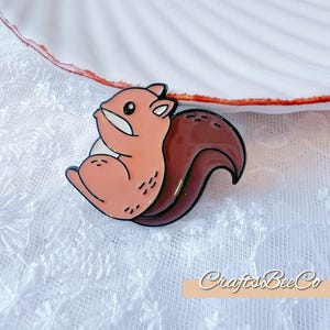 May include: A cartoon squirrel enamel pin with a light pink body, dark brown tail, and black outline. The pin is set against a white lace background and a white seashell-shaped dish with a red rim. The pin has the text "CraftsBeeCo".
