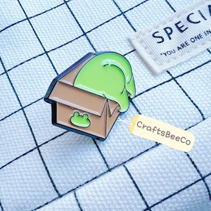 Könnte beinhalten: Ein braun-grüner Emaille-Pin mit einer aus einem Karton springenden Frosch-Darstellung. Der Pin hat einen schwarzen Rand und ein kleines Frosch-Design auf der Box. Ein Etikett mit dem Text "CraftsBeeCo" ist angebracht.