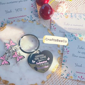 May include: A silver keychain with a heart-shaped charm and butterfly charms. The heart charm has the text "Sometimes you forget you're awesome so this is your reminder". Cards with floral designs and text are also present.