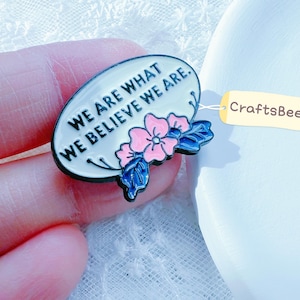 Op de afbeelding: Ovale emaille pin met de tekst "WE ARE WHAT WE BELIEVE WE ARE". De pin heeft een bloemenontwerp met roze en blauwe accenten. Een klein label met de tekst "CraftsBeeCo" is bevestigd.