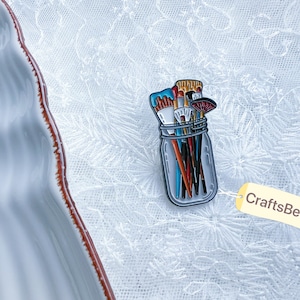 Könnte beinhalten: Ein farbenfroher Emaille-Pin mit einem Einmachglas voller Pinsel. Die Pinsel haben verschiedene Farben, darunter Blau, Orange und Rot. Der Pin hat ein Etikett mit der Aufschrift CraftsBeeCo.