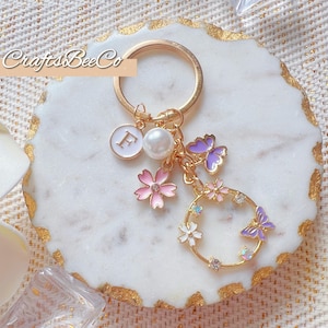 Peut inclure: Porte-clés doré avec des breloques florales et papillons. Le porte-clés comprend un charm initial blanc, une perle, des fleurs roses et un charm circulaire avec des papillons et des cristaux. Le texte "CraftsBeeCo" est visible.
