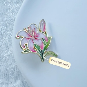 May include: A lily-shaped brooch with white and pink petals, green leaves, and a gold-tone border. The brooch has a tag that reads "CraftsBeeCo".