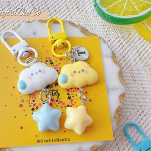 May include: A collection of whimsical keychains featuring cloud and star designs in pastel colors. The keychains have metal clasps and are displayed on a yellow card with star accents. The image includes text that reads "CraftsBeeCo" and "You are my star".
