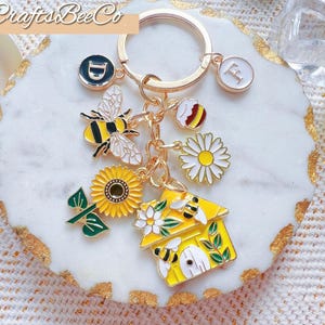 May include: Gold-coloured keyring with bee-themed charms. Charms include a bee, sunflower, beehive, and daisy. The keyring also has a charm with the letter "F" and a charm with a "D" on it. The brand name "CraftsBeeCo" is in the top left corner.