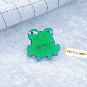 Könnte beinhalten: Eine grüne froschförmige Brosche mit dem Wort „REALLY?“ in der Mitte. Die Brosche hat ein cartoonartiges Design mit großen, ausdrucksstarken Augen. Ein kleines, gelbes Etikett mit dem Text „CraftsBeeCo“ ist angebracht.