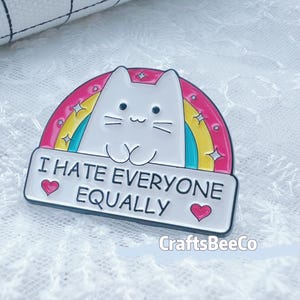 May include: Enamel pin showing a white cat against a rainbow. The pin reads "I HATE EVERYONE EQUALLY" on a white banner, with pink hearts on either side. The pin is set on a white lace surface.