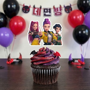 May include: A decorated cupcake with red and purple frosting, bat-shaped decorations, and a cartoon image of three women. The background includes red, black, and purple balloons, and a banner with Korean text.