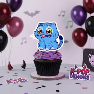 May include: A cupcake topped with a blue cartoon cat with yellow eyes and a pink tongue. The cupcake has purple frosting with sprinkles. The background includes balloons, musical notes, and the text "K-POP DEMONS".