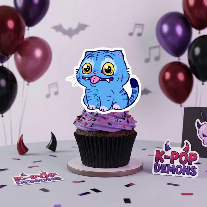 May include: A cupcake topped with a blue cartoon cat with yellow eyes and a pink tongue. The cupcake has purple frosting with sprinkles. The background includes balloons, musical notes, and the text "K-POP DEMONS".