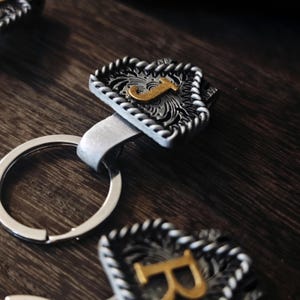 Pode incluir: Um porta-chaves de metal prateado com um pendente retangular decorativo. O pendente apresenta a letra "J" dourada sobre um fundo texturizado e detalhado, emoldurado por uma borda em forma de corda. O porta-chaves está sobre uma superfície de madeira escura.