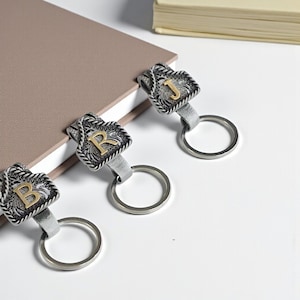 May include: Three silver keychains with decorative rectangular pendants, each featuring a different gold initial: B, R, and J. The pendants have a detailed, textured design and are attached to a silver ring.