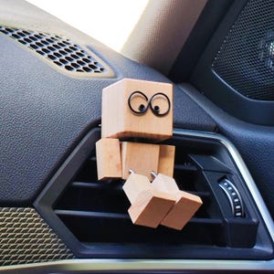 Puede incluir: Un clip de ventilación de coche de madera con forma de pequeño robot. El robot tiene una cabeza cuadrada con ojos de alambre negro y brazos y piernas articulados. El clip está hecho de madera de color claro y está diseñado para sujetarse a una rejilla de ventilación del coche.